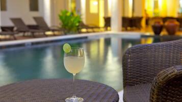 A poolside setting with a glass of a light-colored drink garnished with a lime, wicker furniture, and a pool in the background.