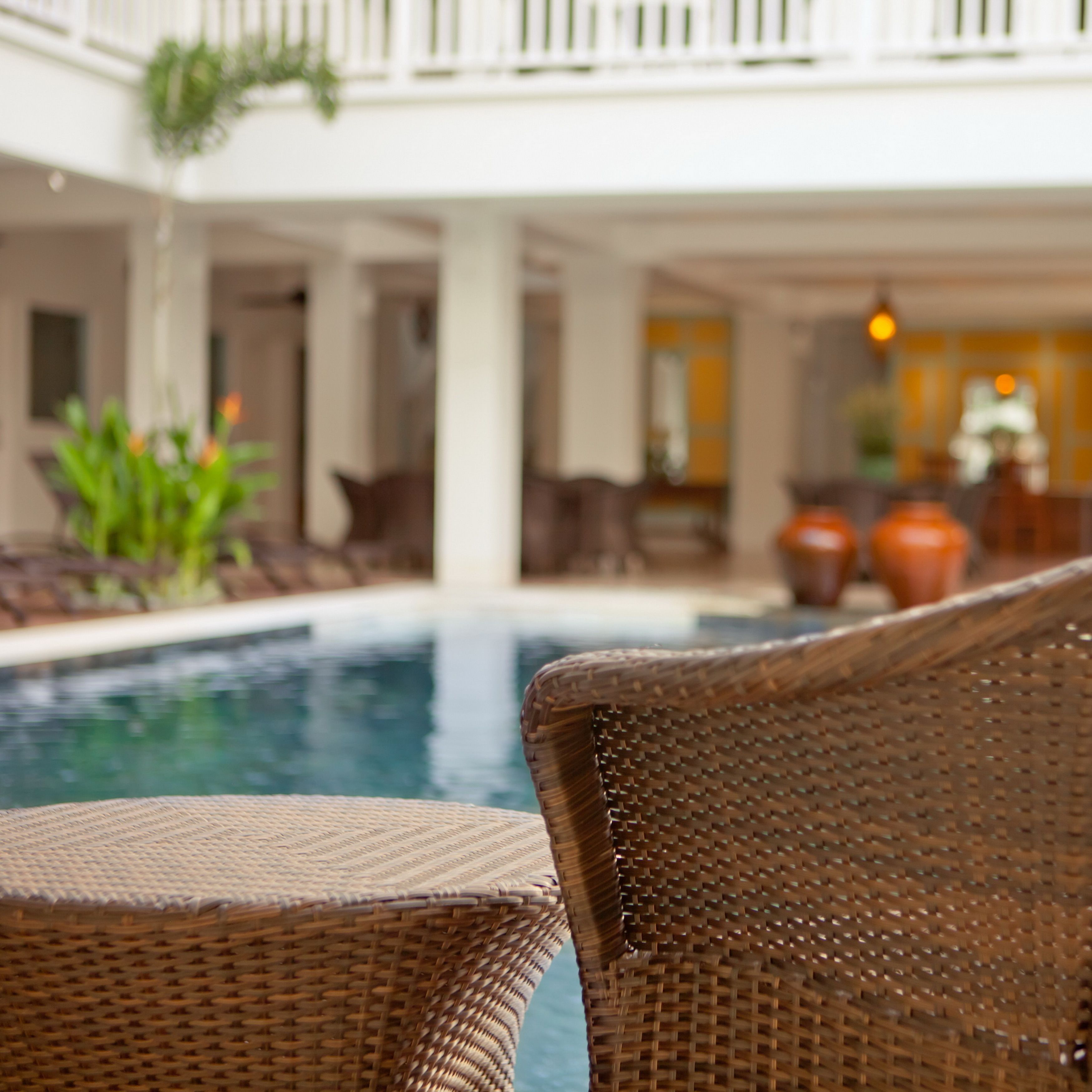 A wicker lounge chair facing a pool with a view of a white building and indoor plants.