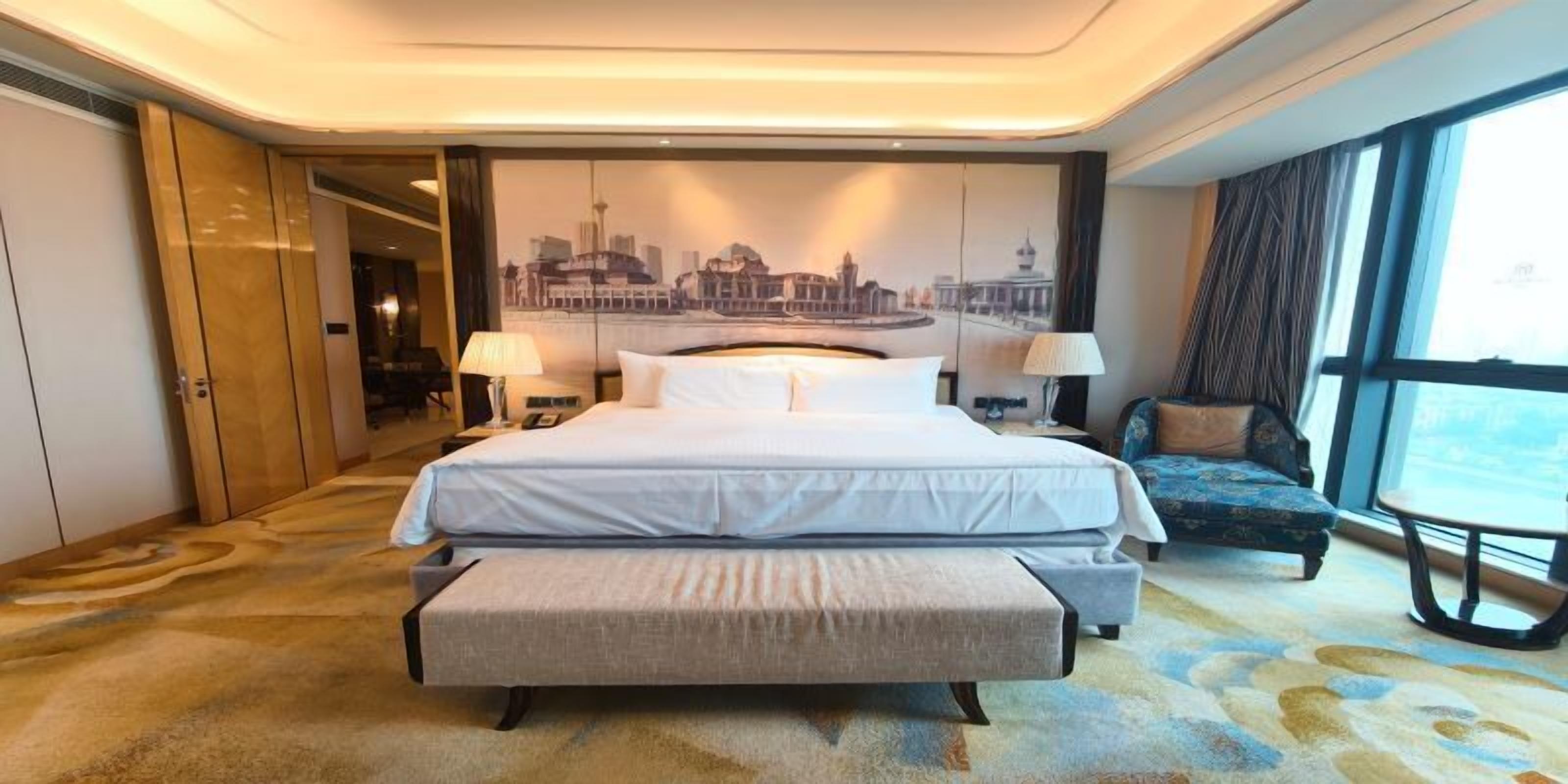 A hotel room with a large bed, a bench, a chair, a desk, and a view of the cityscape.