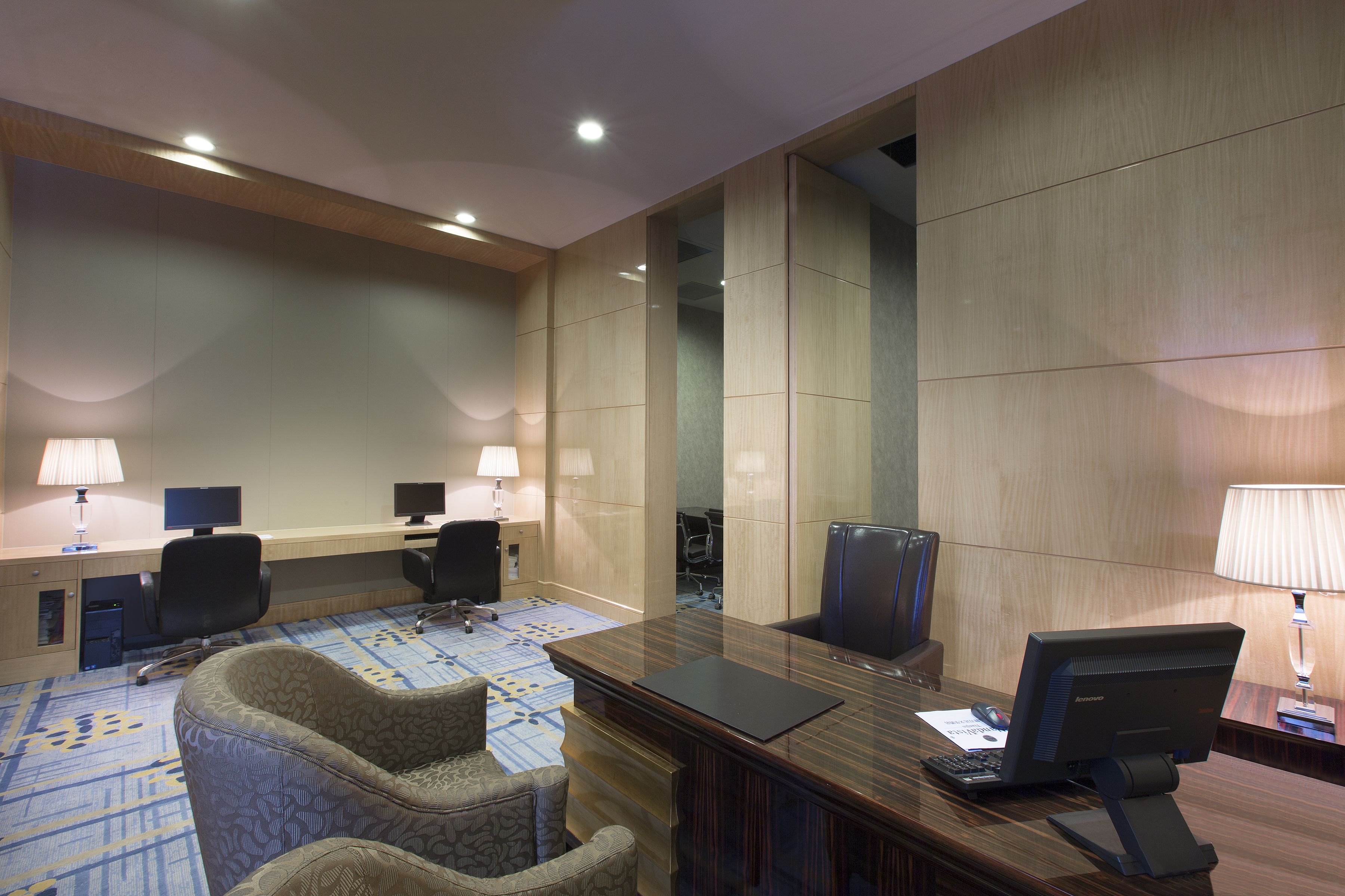 Uma sala de conferências de hotel com uma longa mesa de madeira, duas cadeiras de escritório, uma escrivaninha com um computador e um tapete estampado.
