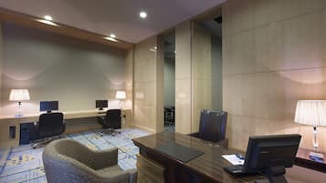 Uma sala de conferências de hotel com uma longa mesa de madeira, duas cadeiras de escritório, uma escrivaninha com um computador e um tapete estampado.