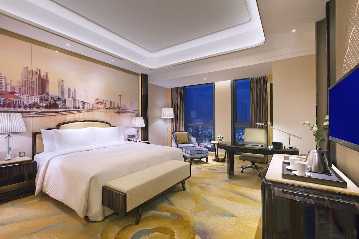 Une chambre d’hôtel avec un grand lit, un bureau avec une chaise, une télévision et une fresque murale représentant un paysage urbain.