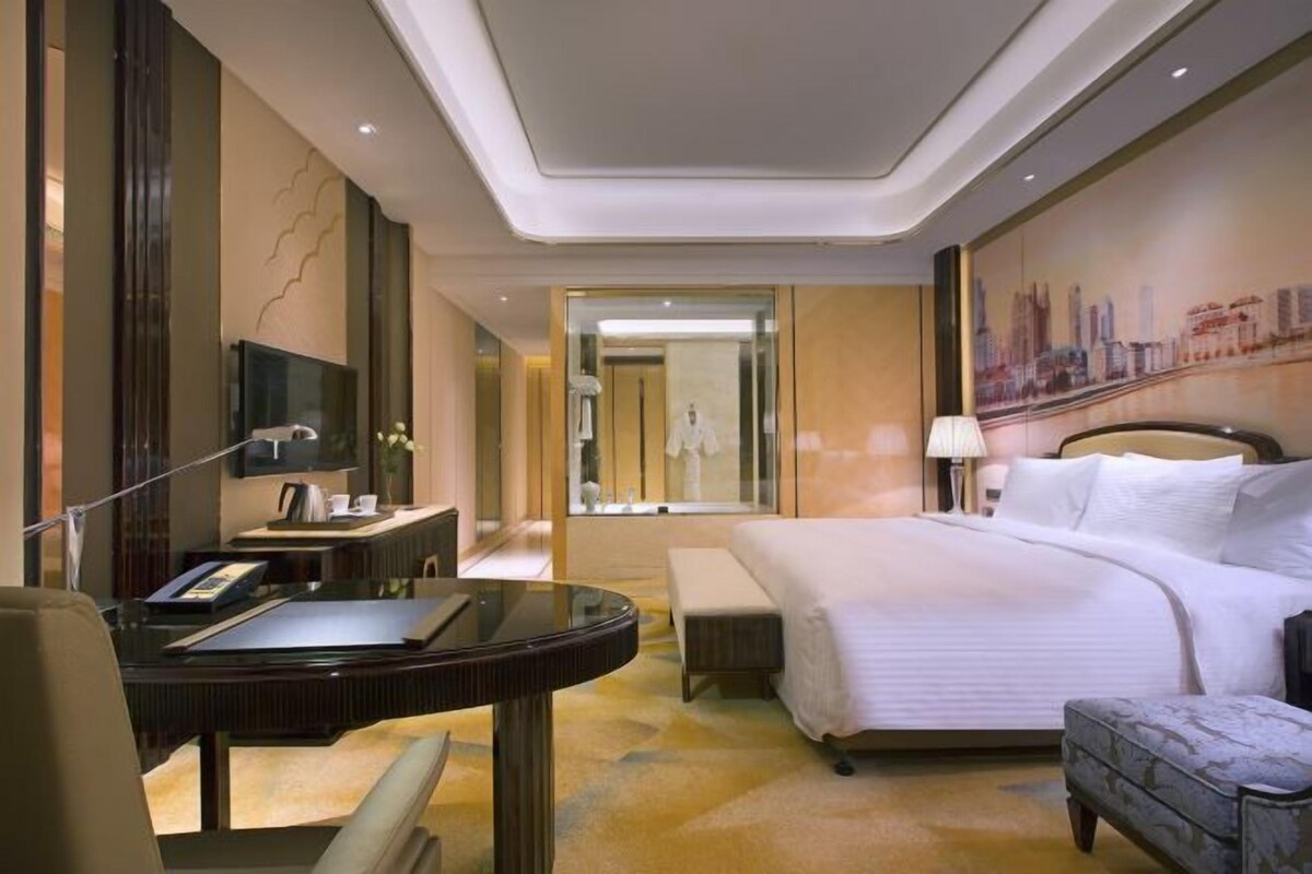 Habitación de hotel con cama, escritorio, silla, televisión y baño visible a través de una puerta de vidrio.