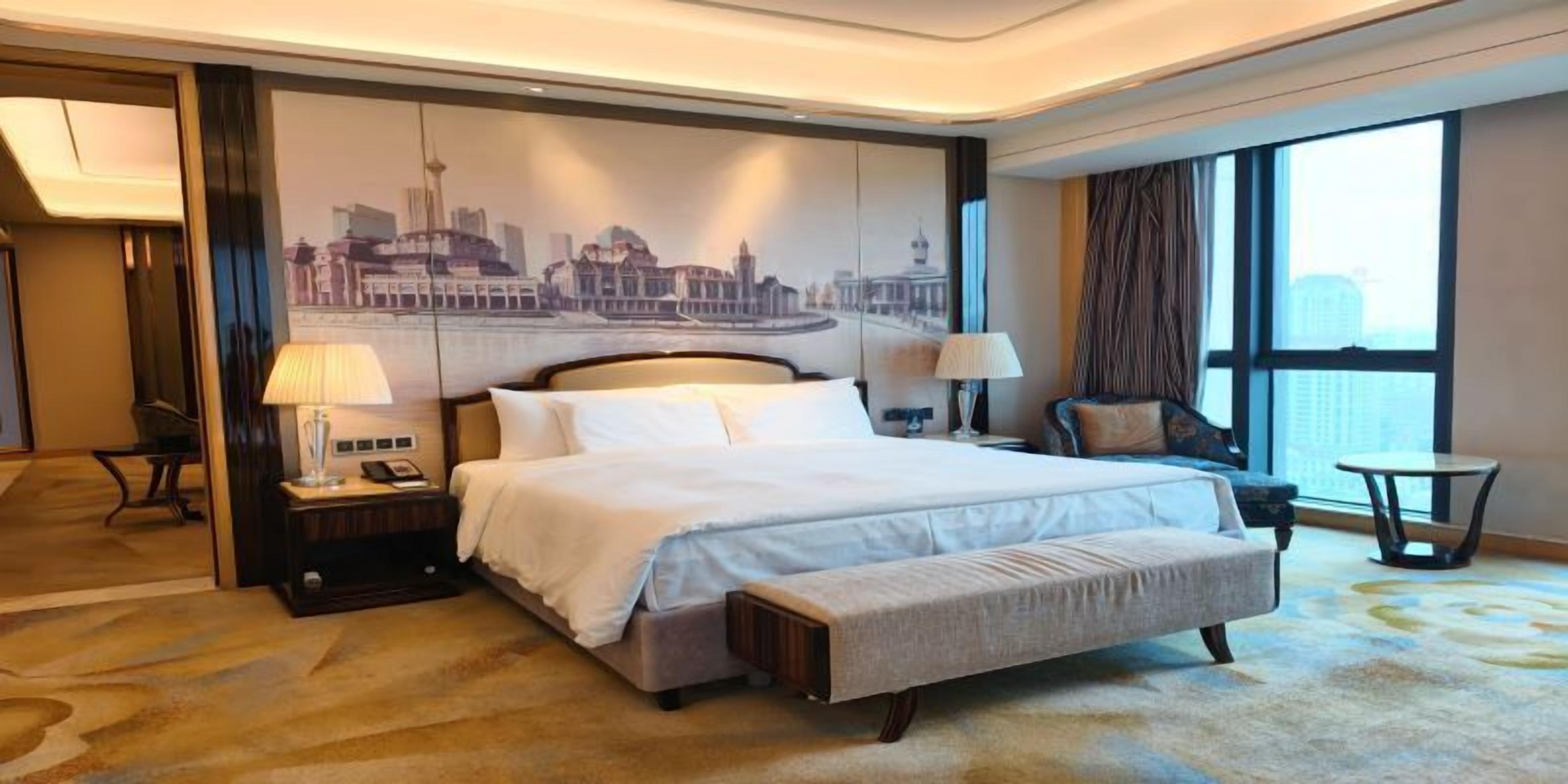 Una habitación de hotel con una cama grande, un mural de paisaje urbano, un escritorio con lámpara, una silla y una ventana con cortinas.