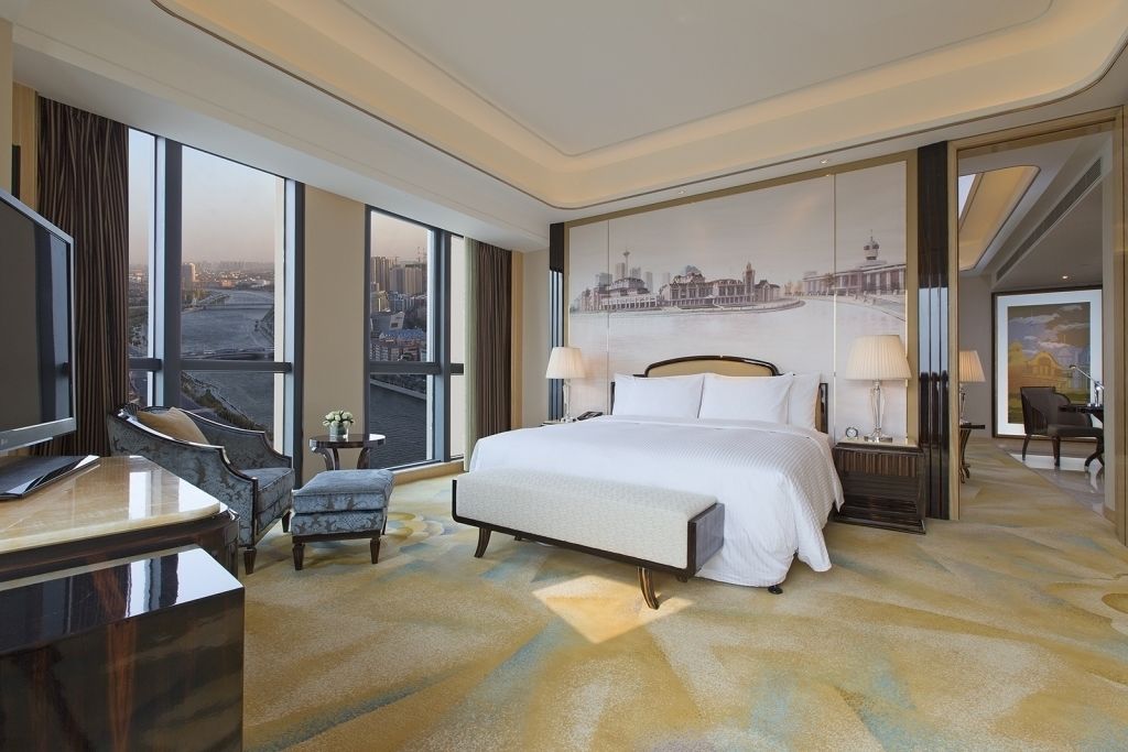 A hotel room with a large bed, a desk, and a chair. There is a large window with a view of a cityscape.