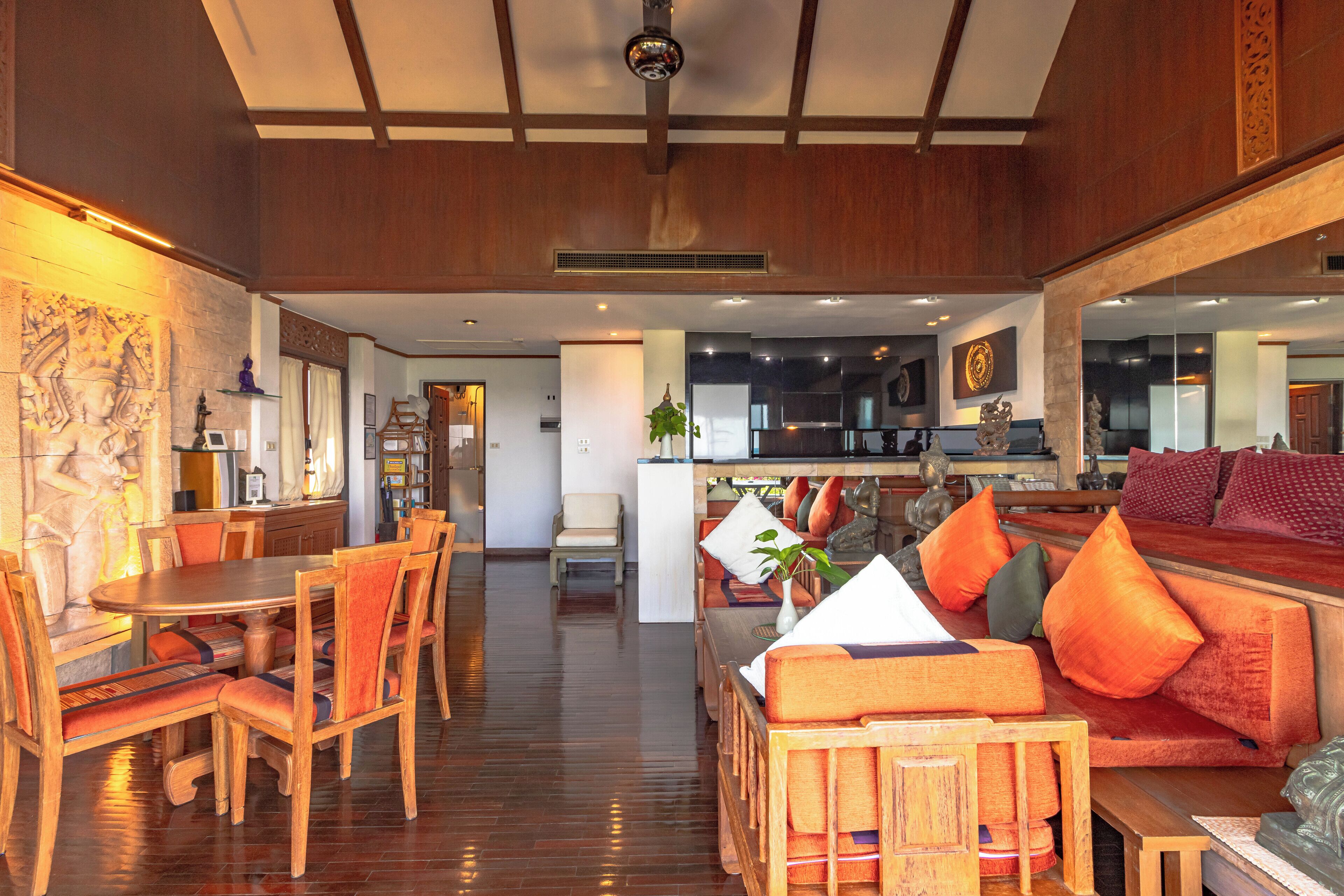 A spacious room with a wooden dining table and chairs, a sofa with orange cushions, and a kitchen area in the background.