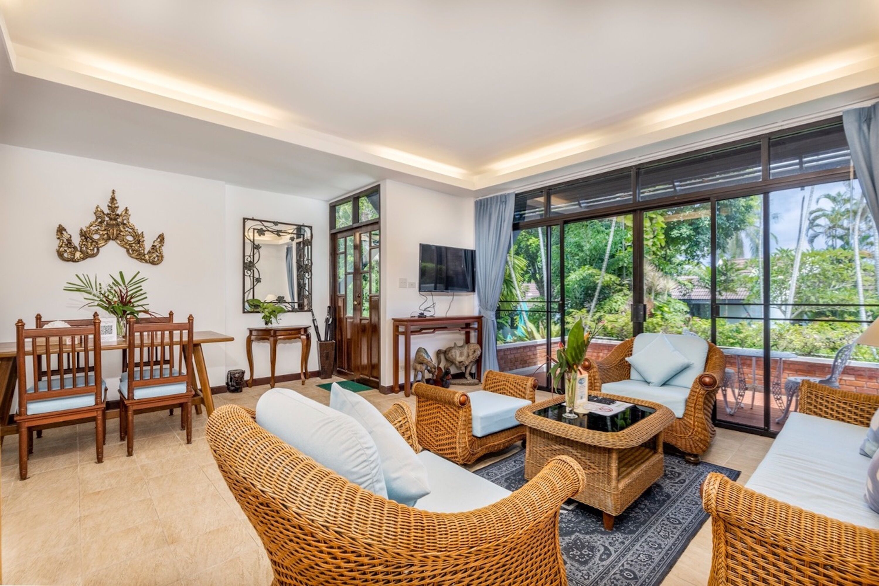 A spacious living area with wicker furniture, a dining table, and a large window offering a view of greenery.