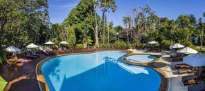 Una amplia piscina exterior con tumbonas y sombrillas, rodeada de exuberante vegetación y altos árboles.