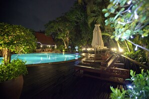 Un área de piscina nocturna con maderas, sillones de descanso y sombrillas.