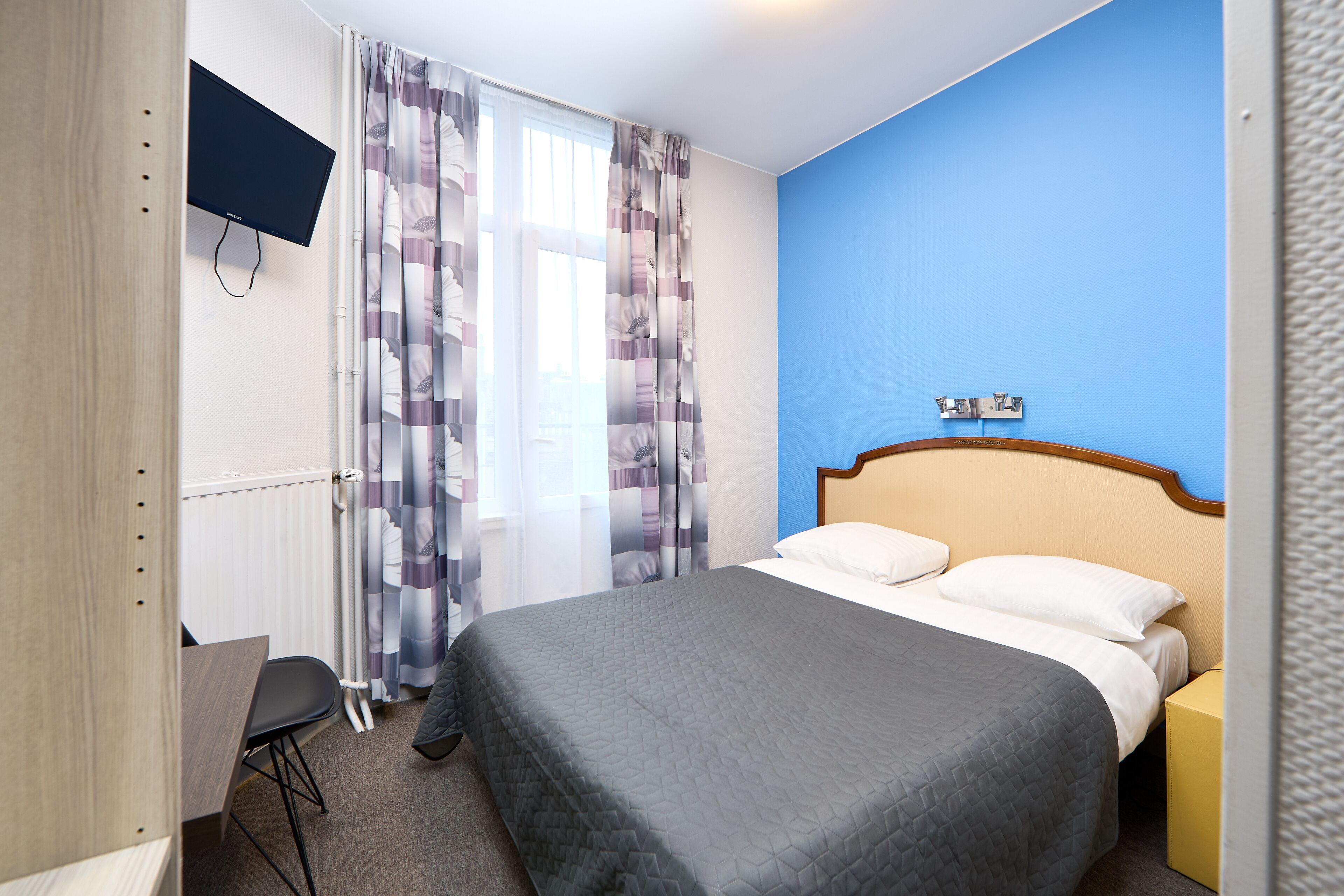Een hotelkamer met een bed, een bureau, een aan de muur bevestigde televisie en een raam met gordijnen.