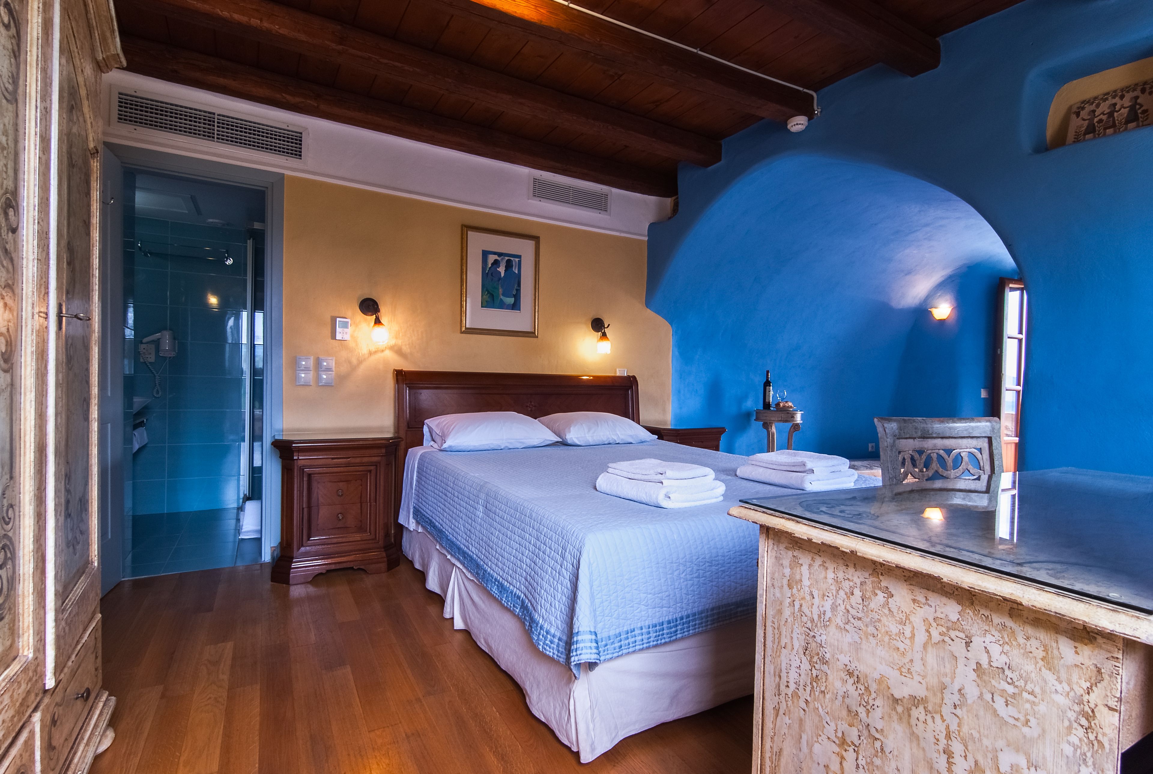 Un dormitorio con techo de madera, una cama con colcha azul, una mesita de noche con lámpara y un baño visible a través de una puerta abierta.