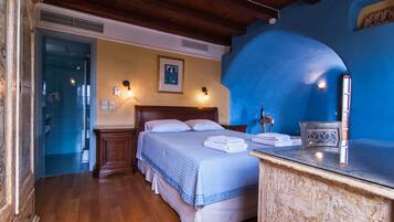 Un dormitorio con techo de madera, una cama con colcha azul, una mesita de noche con lámpara y un baño visible a través de una puerta abierta.