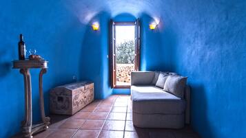 Una habitación con pared azul, una mesa de madera con una botella y un vaso, un sofá y vistas a través de una puerta abierta.