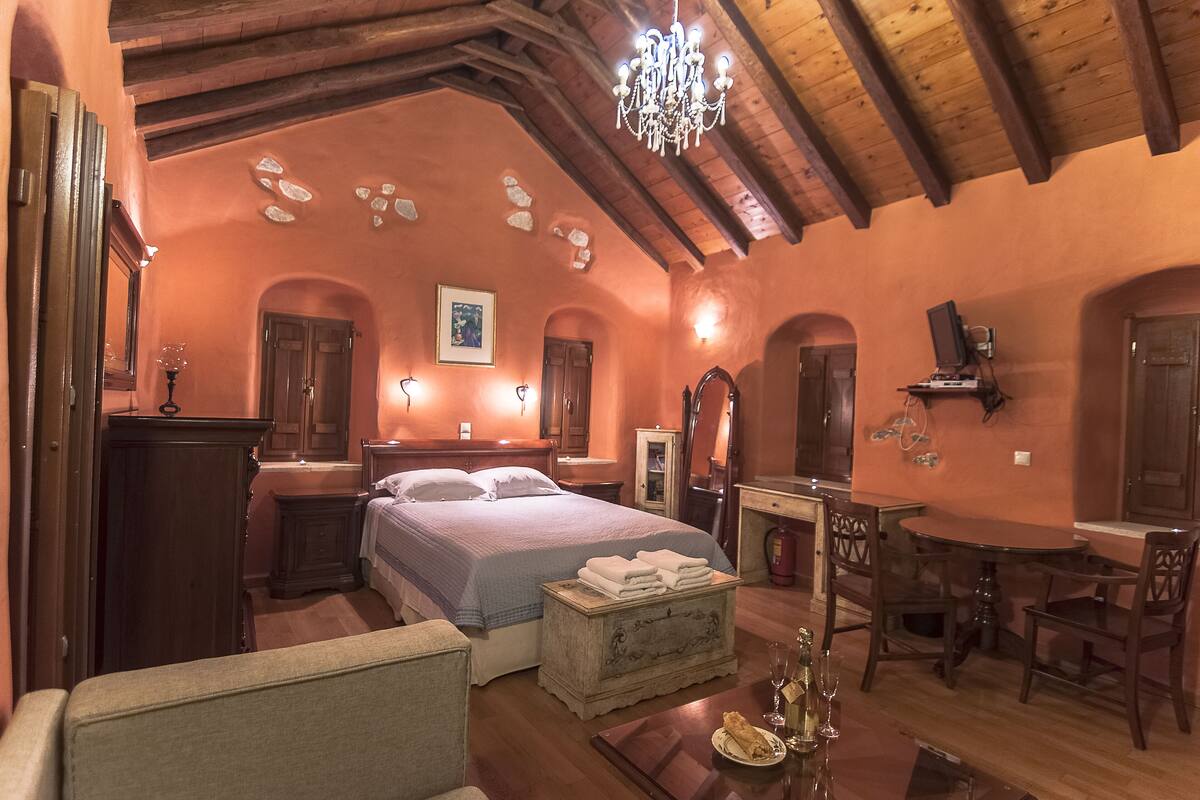 Quarto com cama, mesas de cabeceira, cômoda, lustre e um espaço de jantar com cadeiras e mesa.