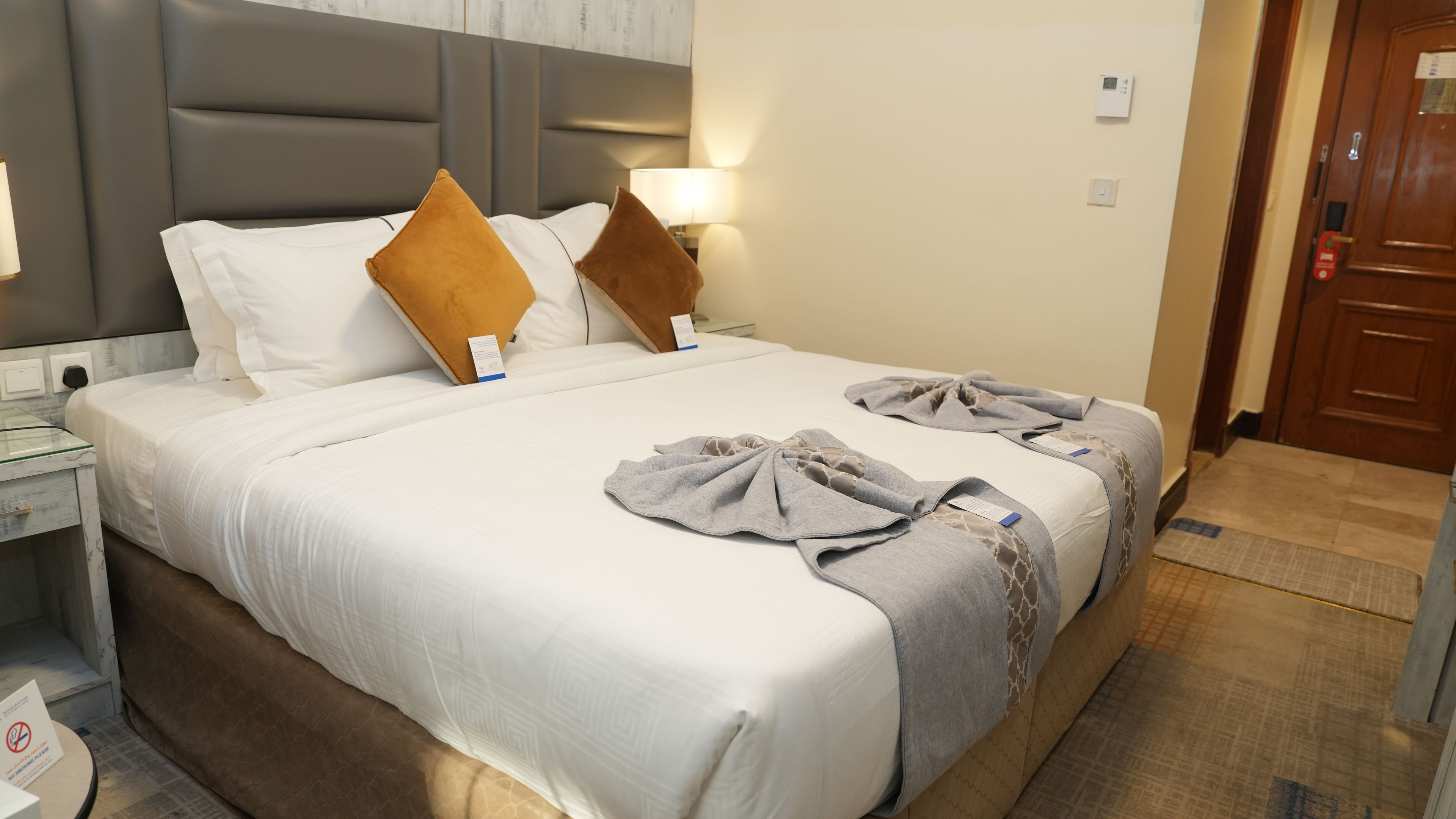 A hotel room with a large bed, two pillows, a bedside table, a nightstand, a lamp, a door, and a wall-mounted thermostat.