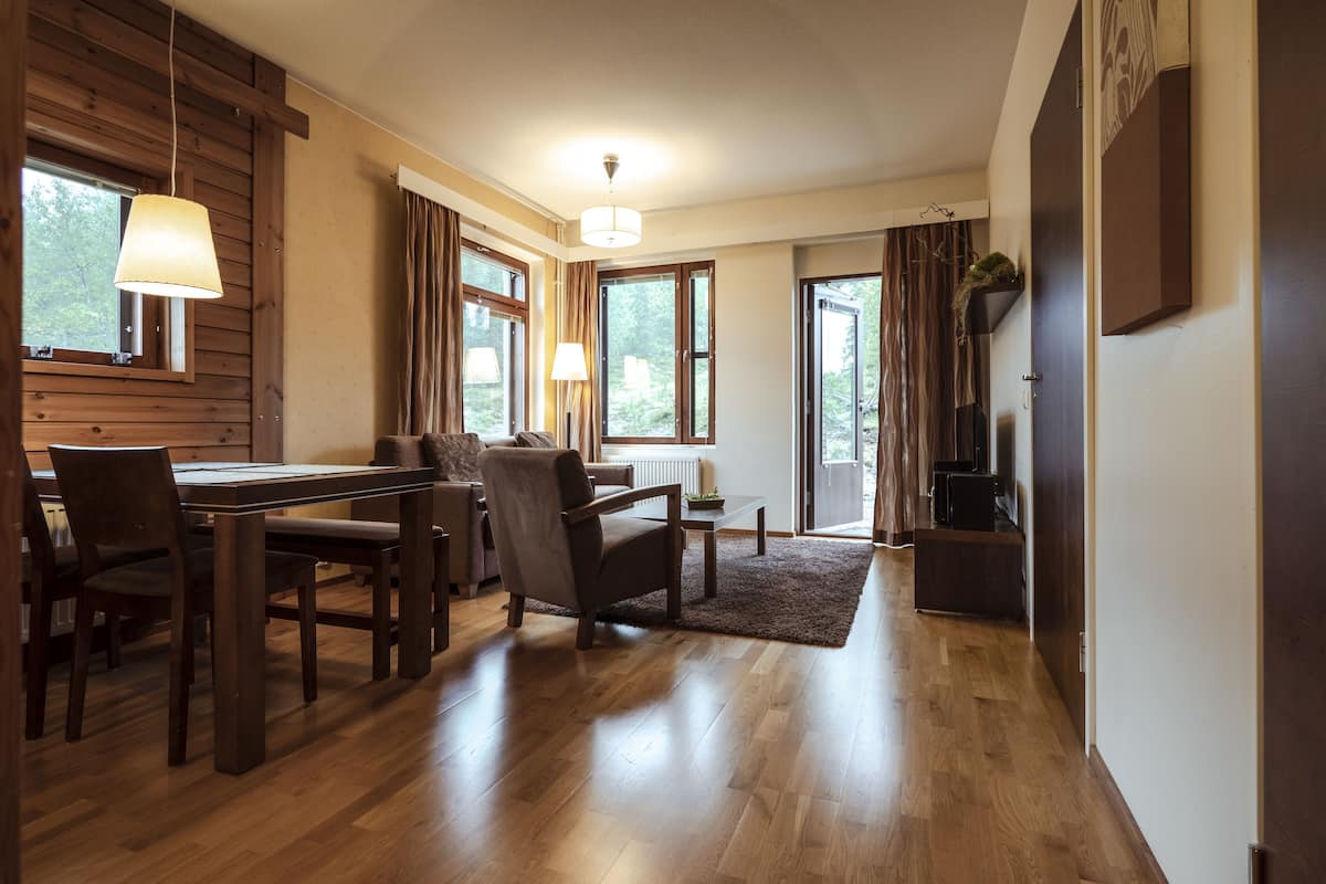 A spacious living area with a wooden dining table, chairs, a sofa, and a coffee table. There are large windows with curtains, a hanging light fixture, and a door leading to another room.