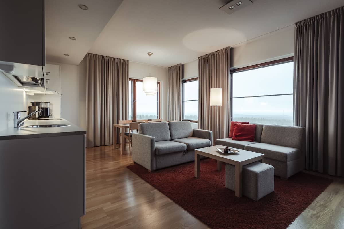 A modern hotel room with a kitchenette, a grey sofa set, a red rug, and large windows with curtains.