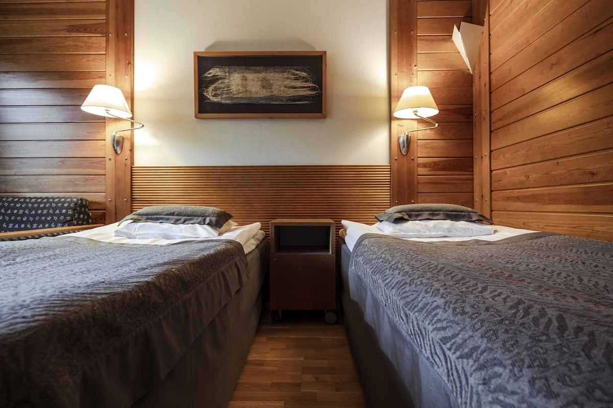 A room with two beds, wooden paneling, a small table, and wall-mounted lamps.