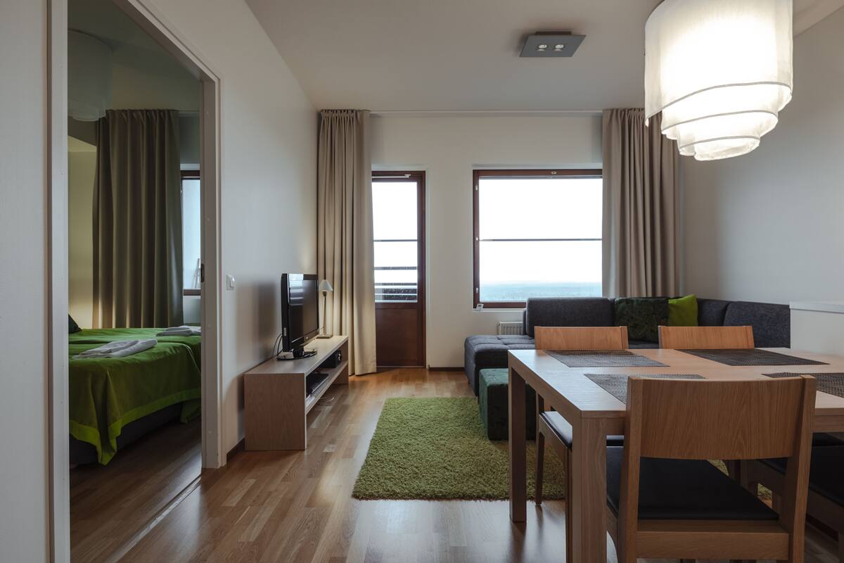 A modern hotel room with a wooden floor, a green bed, a television, a sofa, a dining table with chairs, and a large window providing a view of the sea.