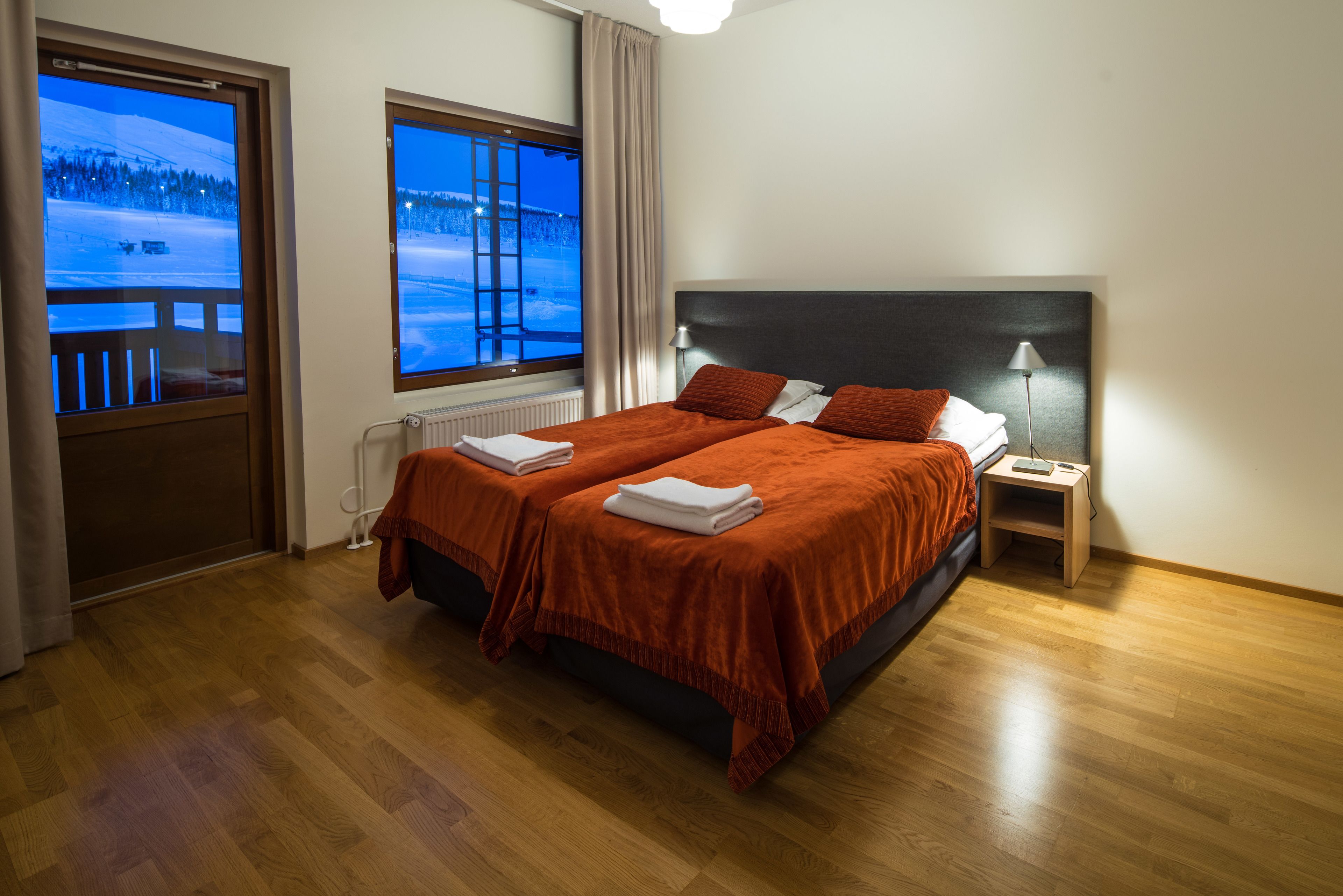 A bedroom with a large bed, two bedside tables, a window with a view of a snowy landscape, and a wooden floor.