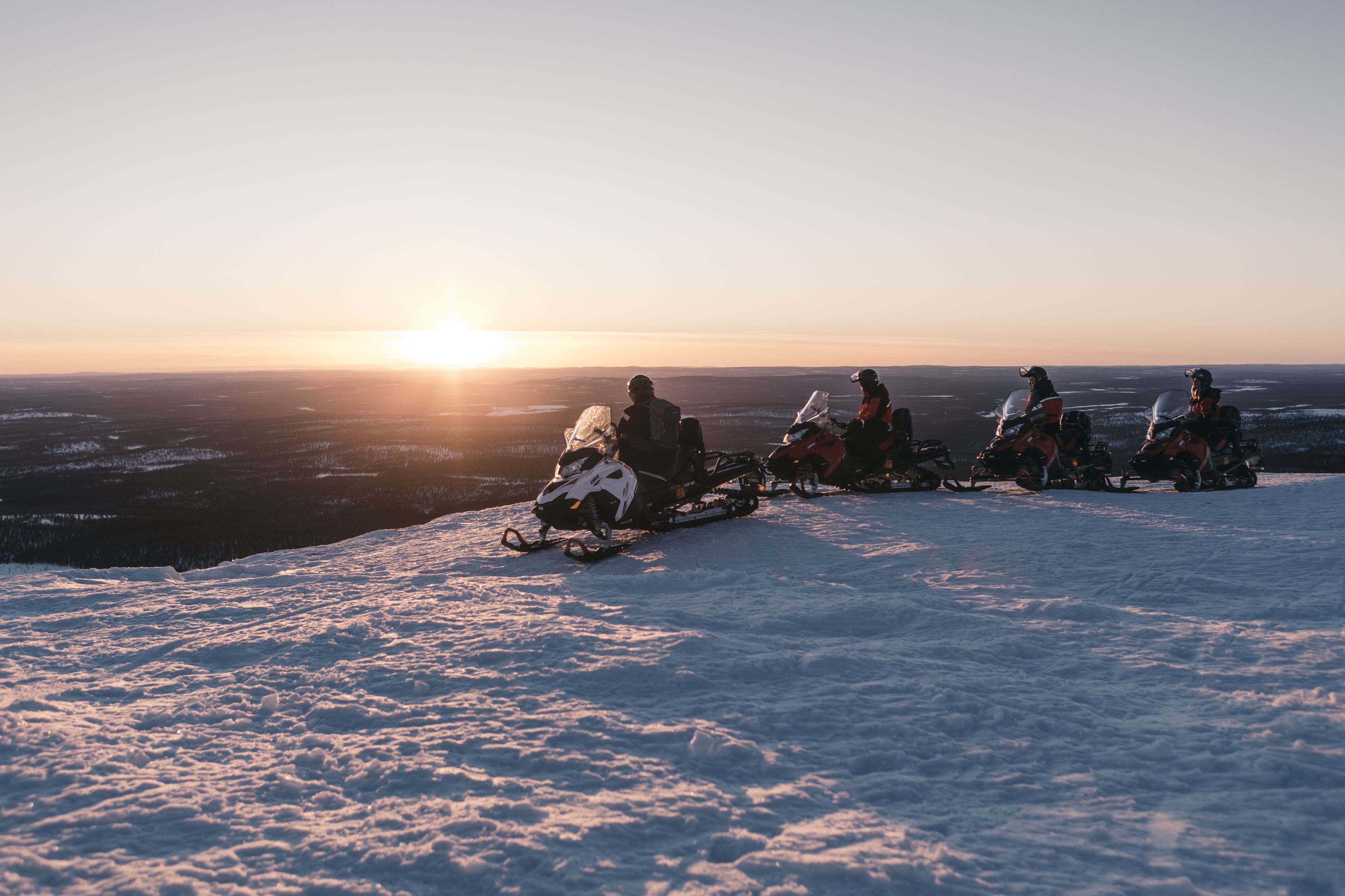Un groupe de personnes sur des motoneiges, évoluant dans un paysage enneigé, avec un coucher de soleil en arrière-plan.