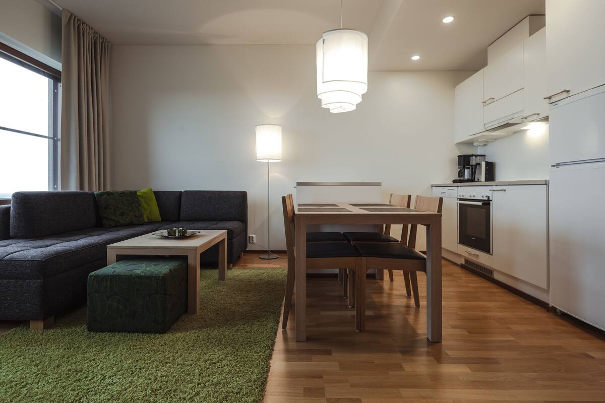 A modern living room with a sofa, ottoman, and a dining table with chairs. There is a kitchen area with cabinets and a coffee machine.
