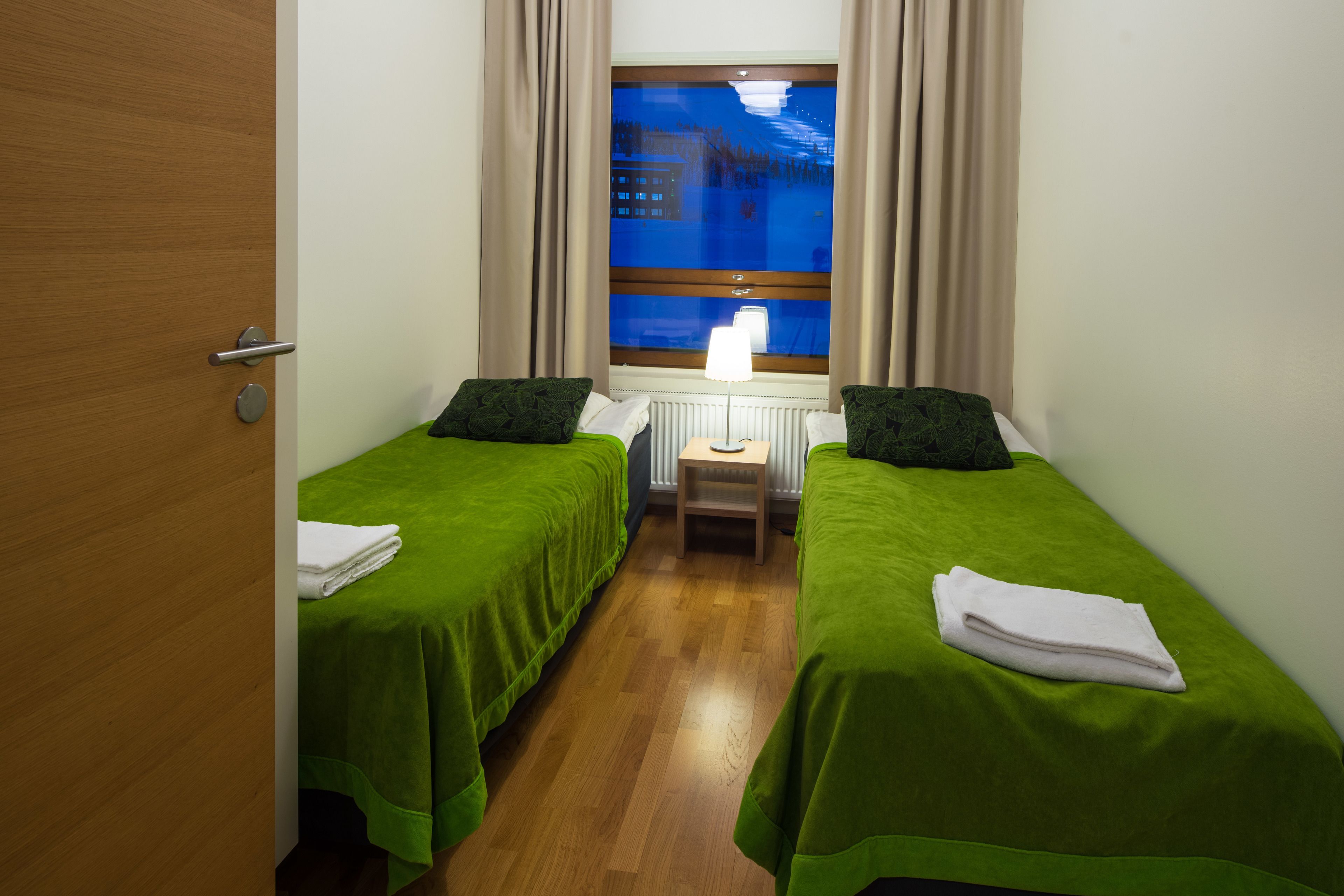 A room with two beds, a window with a view of a cityscape, a radiator, and a door.