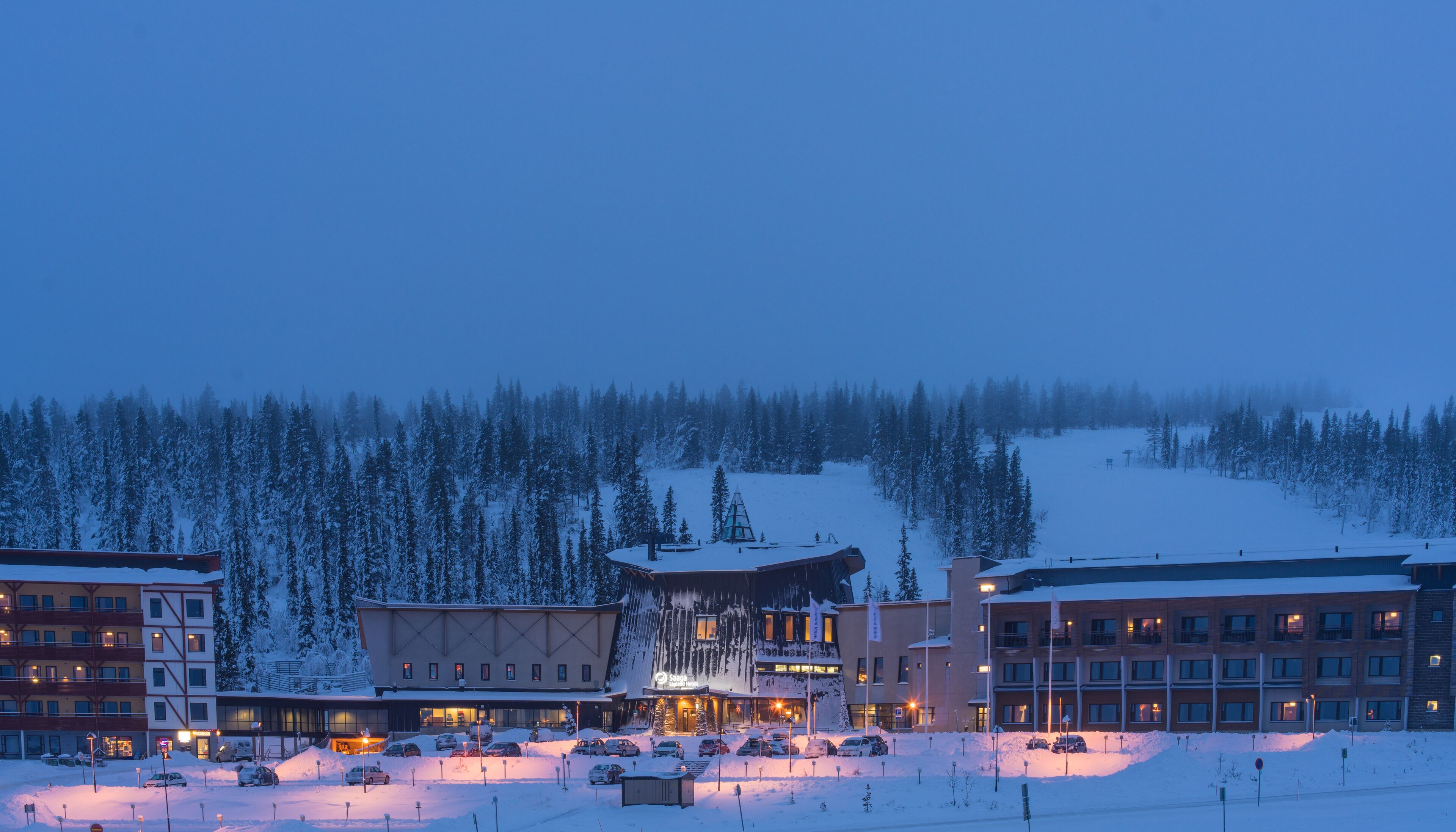 Ett hotell med en huvudbyggnad, flera parkeringsplatser och en snötäckt skog i bakgrunden.