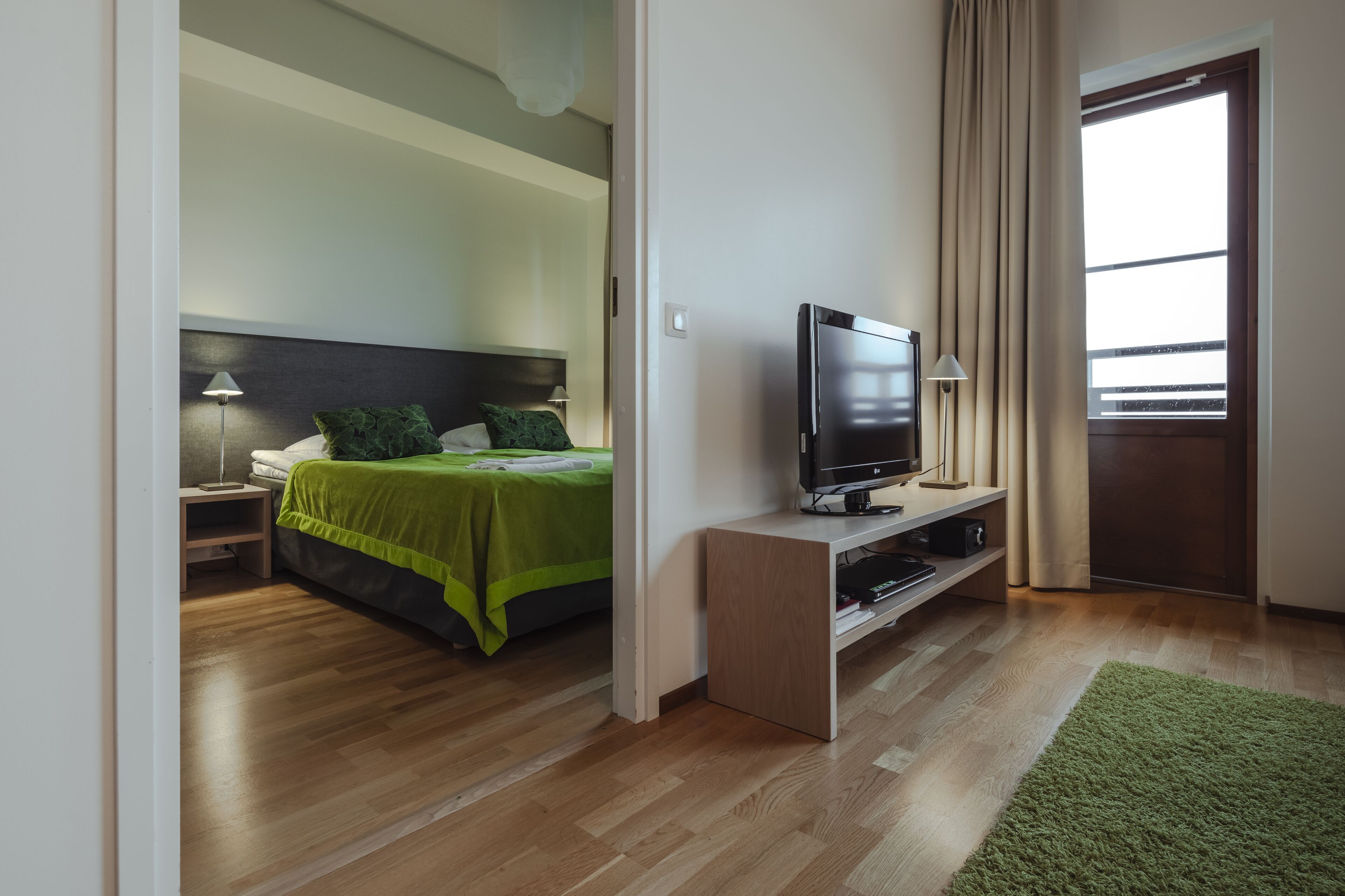 A modern hotel room with a bed, a TV on a stand, a nightstand, and a window with curtains.