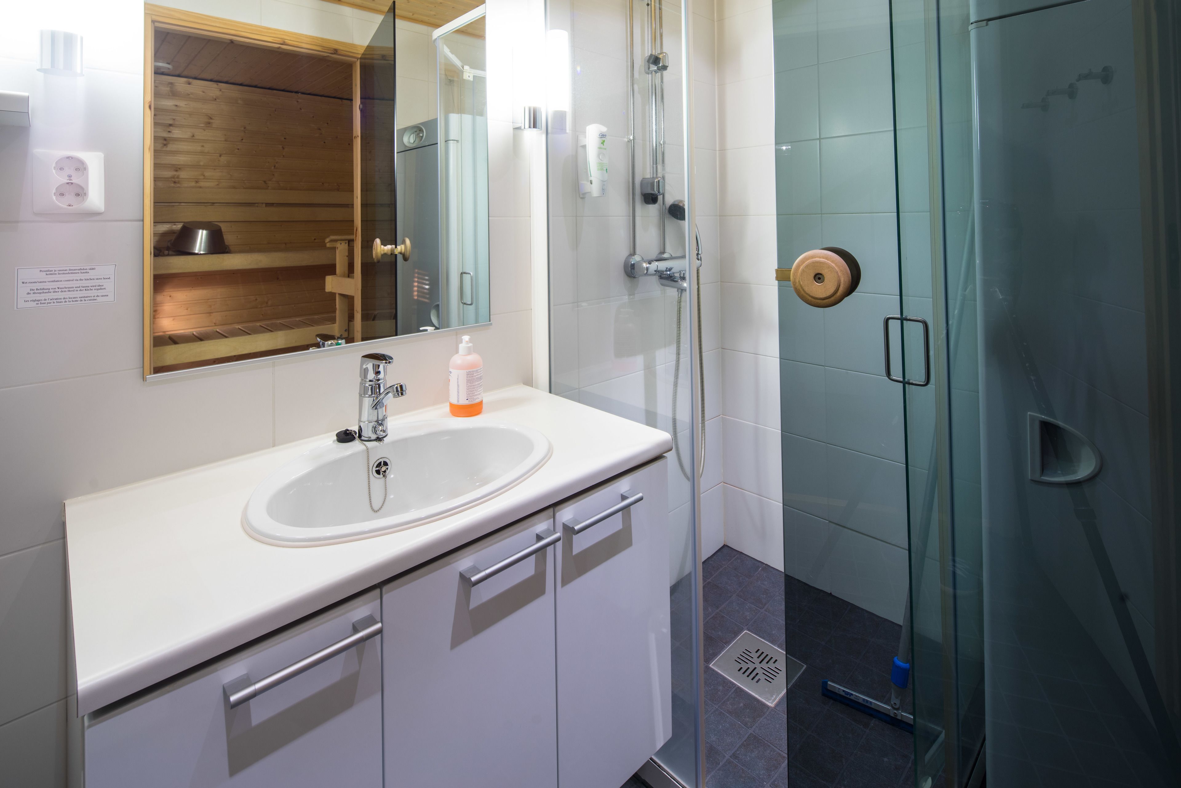 Ein modernes Badezimmer mit einem weißen Waschbecken, einem Spiegel, einer Duschkabine aus Glas und einem Holzregal.