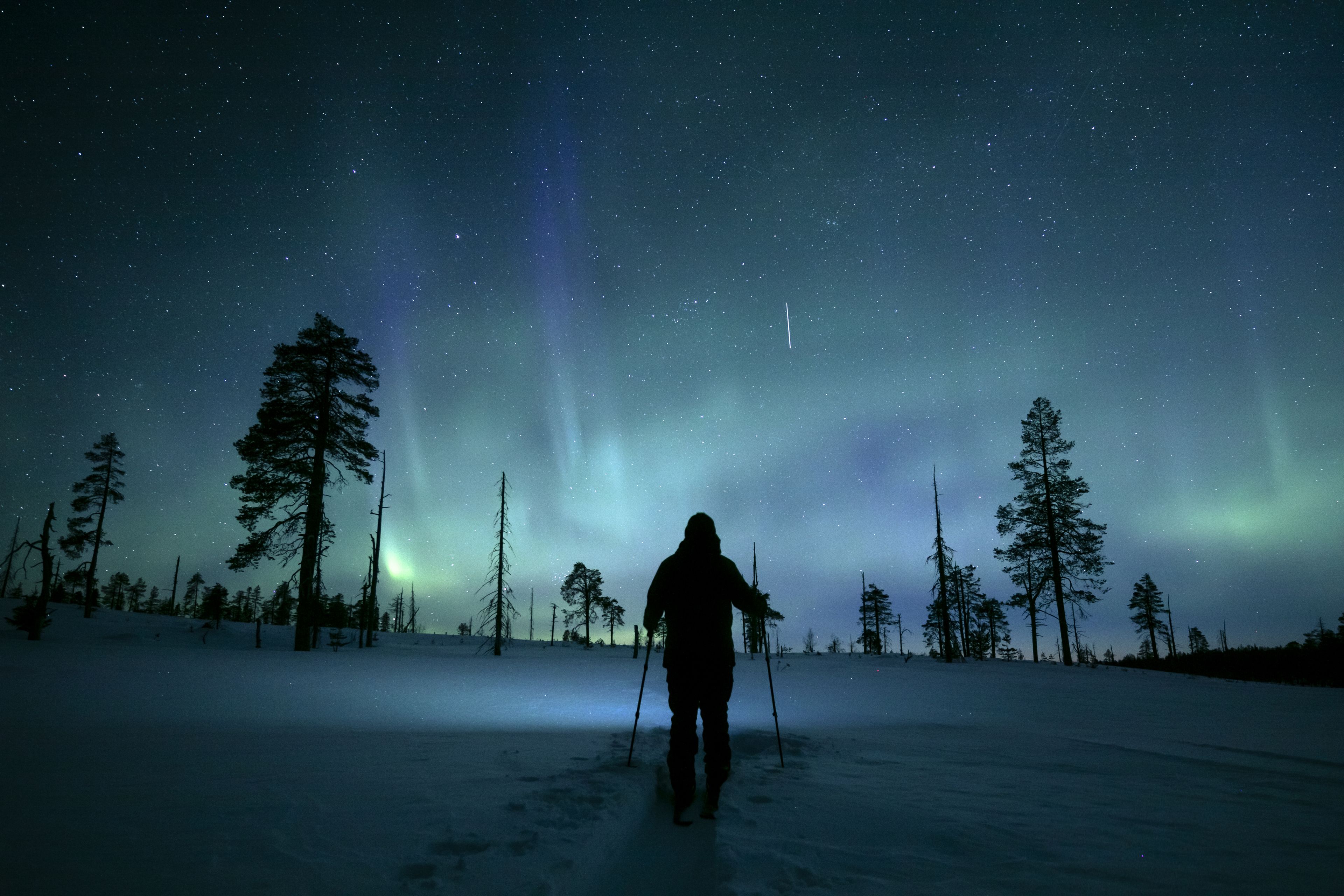 A person walking in a snowy landscape under a starry night sky with visible aurora borealis.