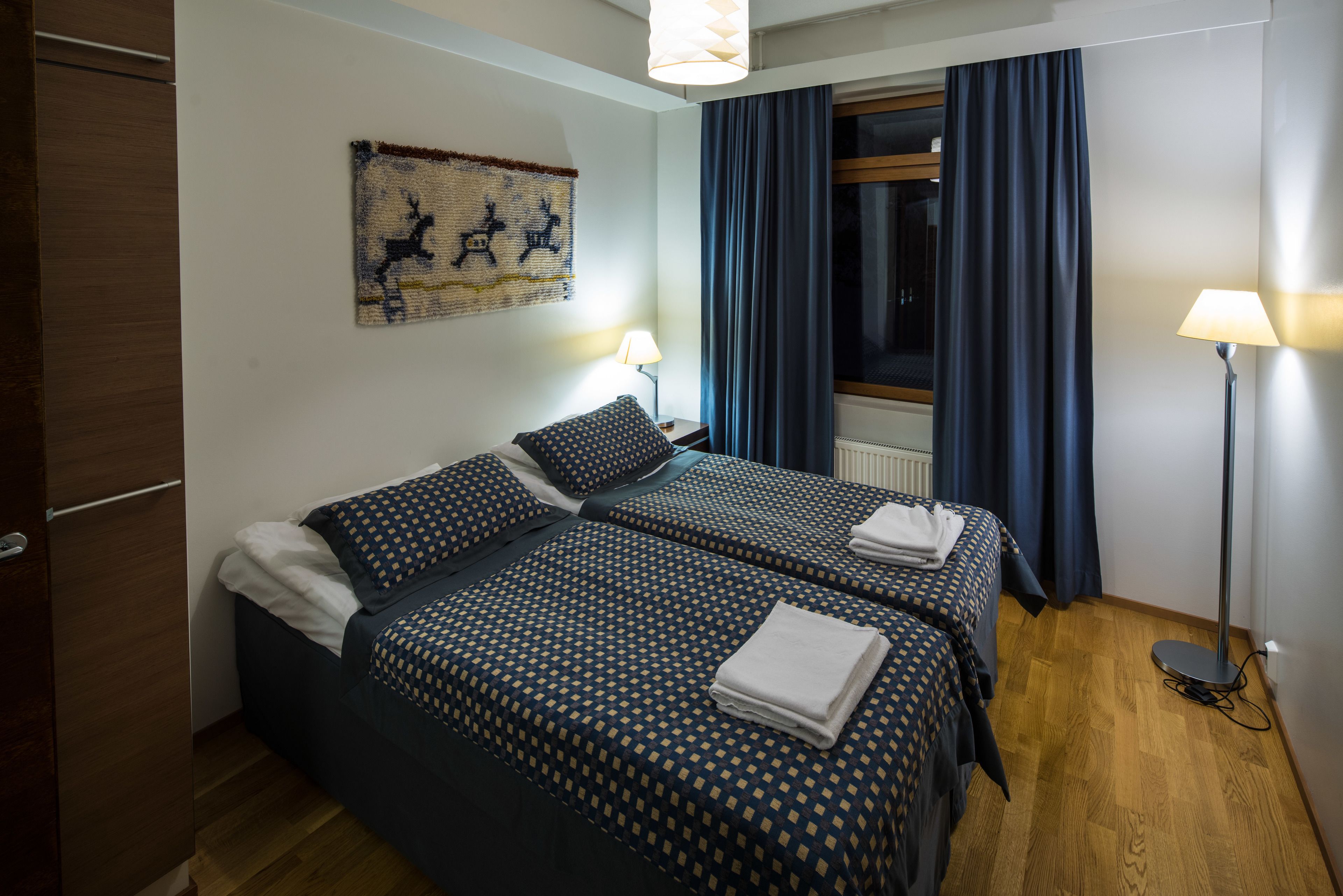 A hotel room with two beds, a wooden wardrobe, a wall art piece, a window with curtains, and two bedside lamps.