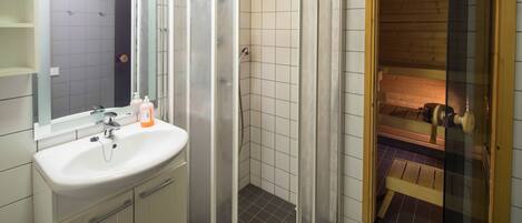 Un baño moderno con ducha envuelta en cristal, un lavabo con artículos de aseo y una sauna de madera visible a través del vidrio.