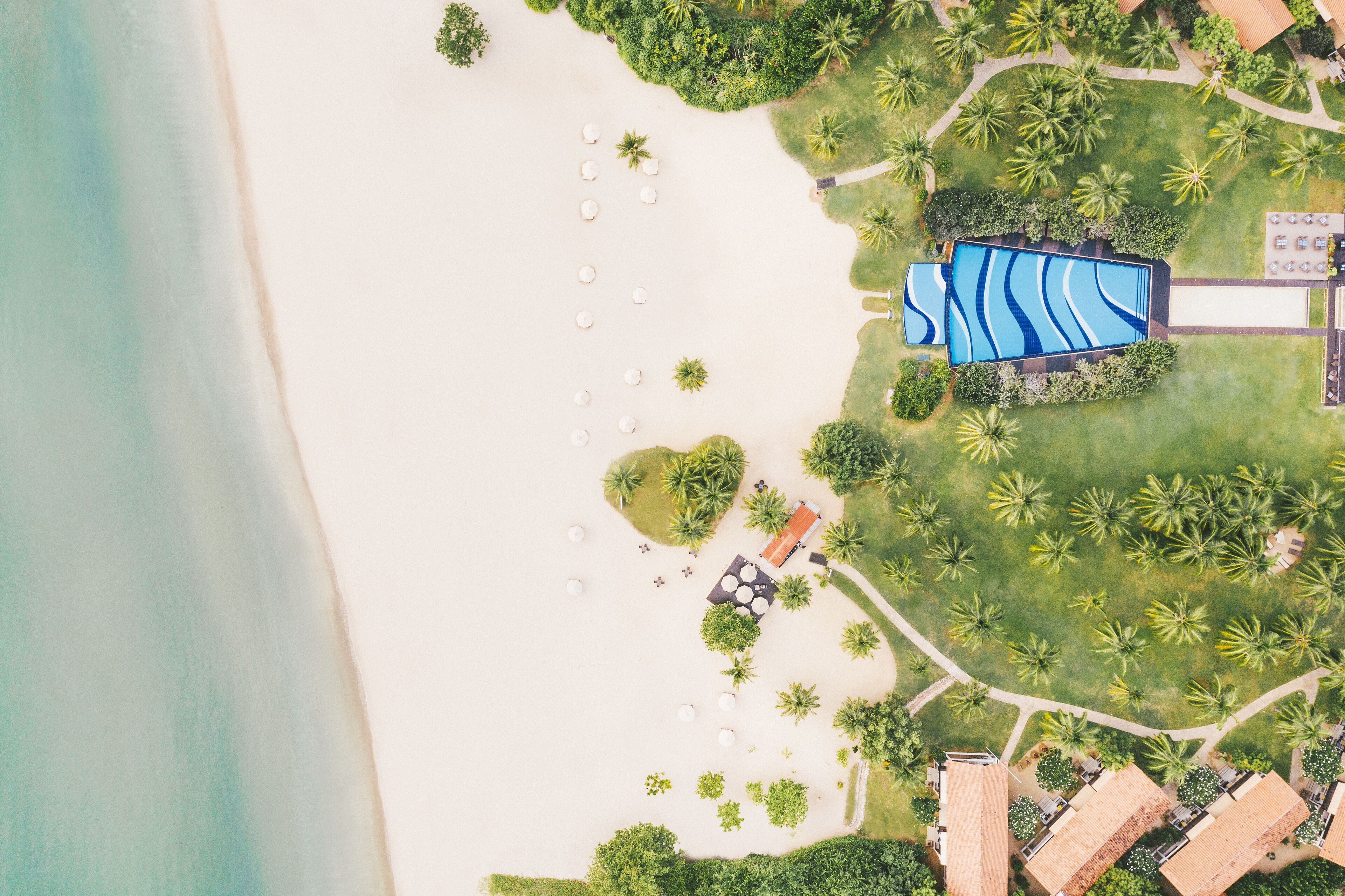 An aerial view of a resort with a swimming pool, beach umbrellas, and palm trees.