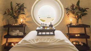 Um quarto de spa com uma grande janela circular, uma mesa de massagem e duas mesinhas laterais com velas e plantas.