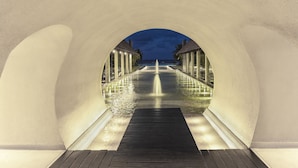 Um corredor com um elemento de água ao centro, ladeado por edifícios com colunas iluminadas.