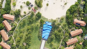 Vista aérea de um resort com vários edifícios, piscina com design de ondas azuis e área de praia com guarda-sóis e palmeiras.