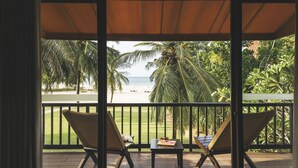 Uma área de estar coberta ao ar livre, com duas cadeiras e uma mesa, com vista para uma praia com palmeiras e o oceano.
