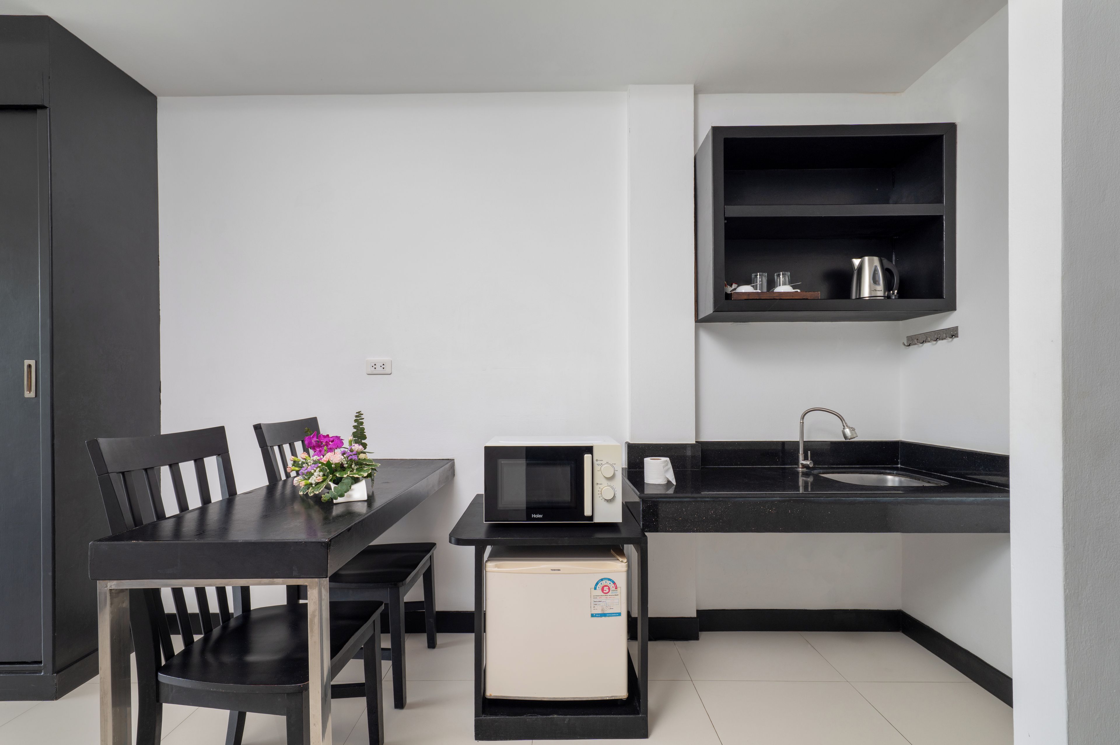 Cocina compacta con una mesa de comedor, sillas, microondas, refrigerador pequeño, pileta y estante con utensilios de cocina.