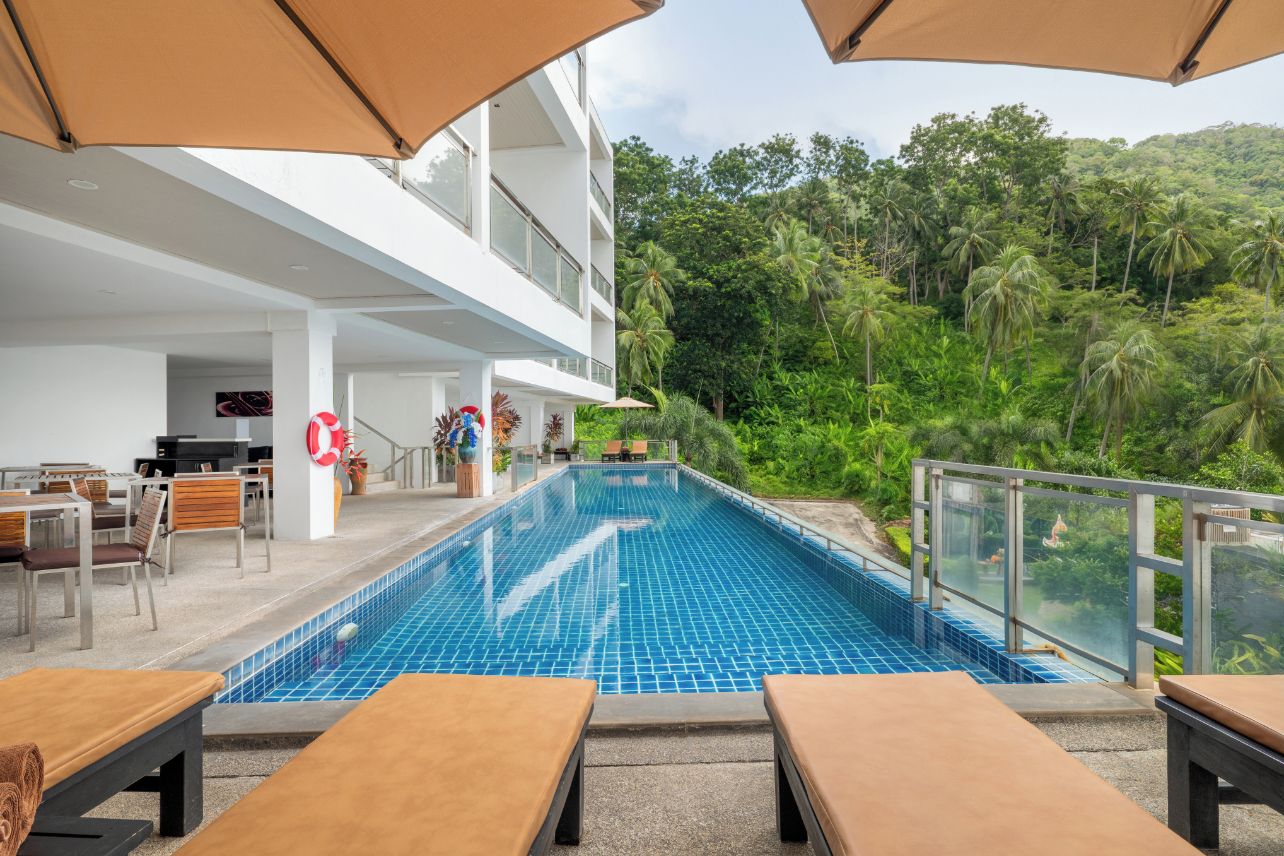 Una piscina con sillones y sombrillas, rodeada de vegetación exuberante y un edificio de varios pisos.