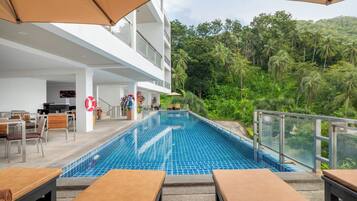 Una piscina con sillones y sombrillas, rodeada de vegetación exuberante y un edificio de varios pisos.