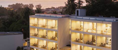 A multi-story hotel with balconies, a pool area, and a view of the sea at sunset.