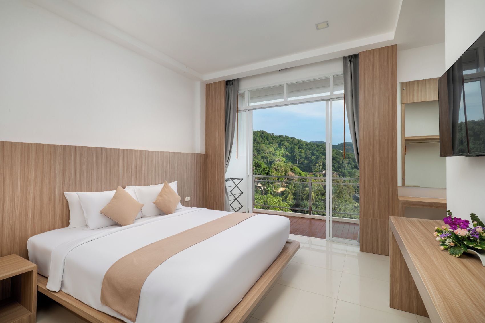 Habitación de hotel con una cama grande, cabecera de madera, balcón con vista a zonas verdes y televisor de pantalla plana montado en la pared.