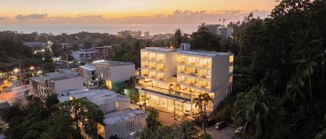 A multi-story hotel with a modern design, surrounded by lush greenery and a view of the sea at sunset.