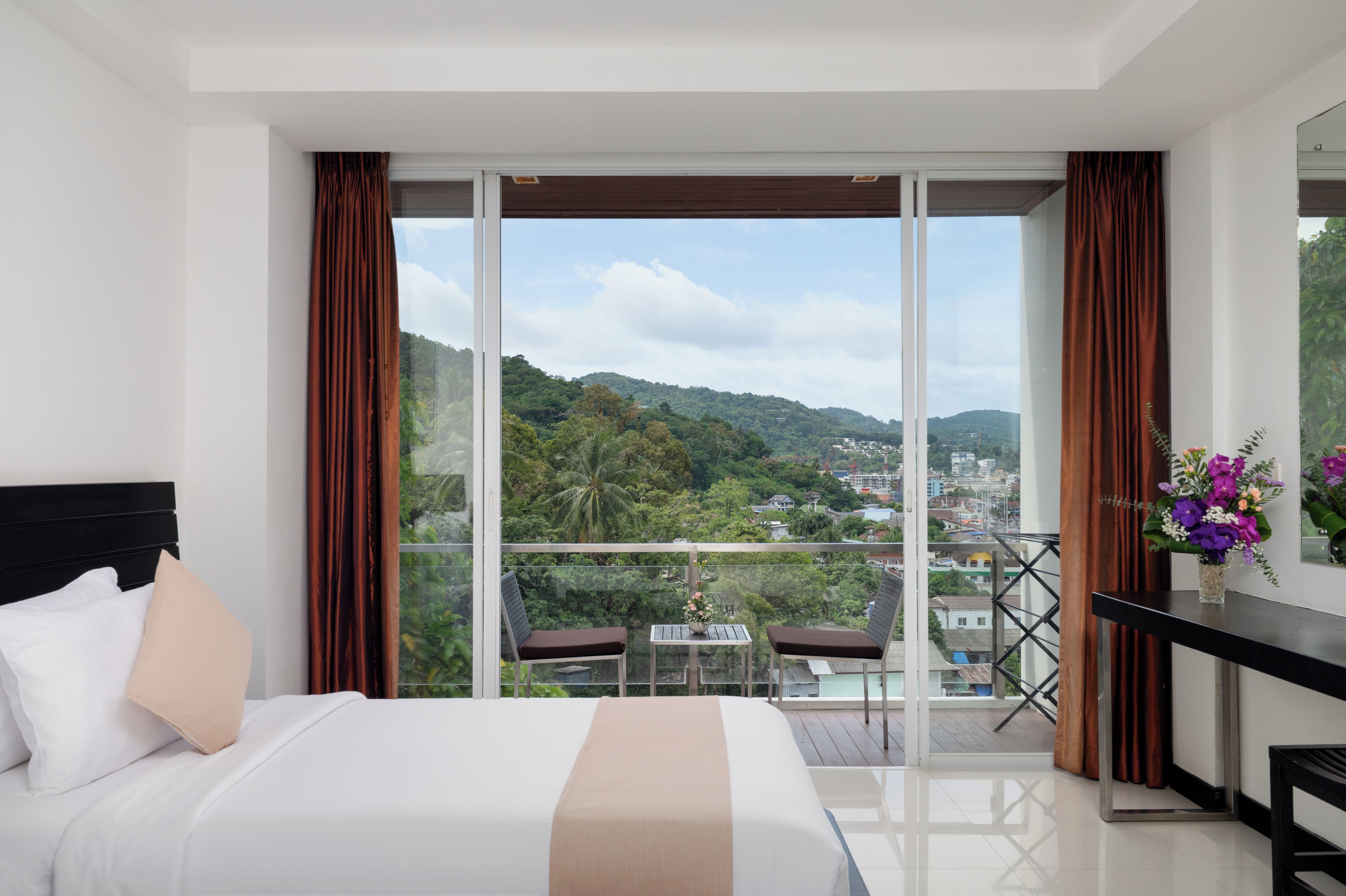 Quarto de hotel com cama, uma pequena mesa com um vaso de flores e uma varanda com vista para a cidade e colinas.