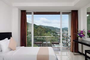 A hotel room with a bed, a small table with a vase of flowers, and a balcony with a view of a city and hills.