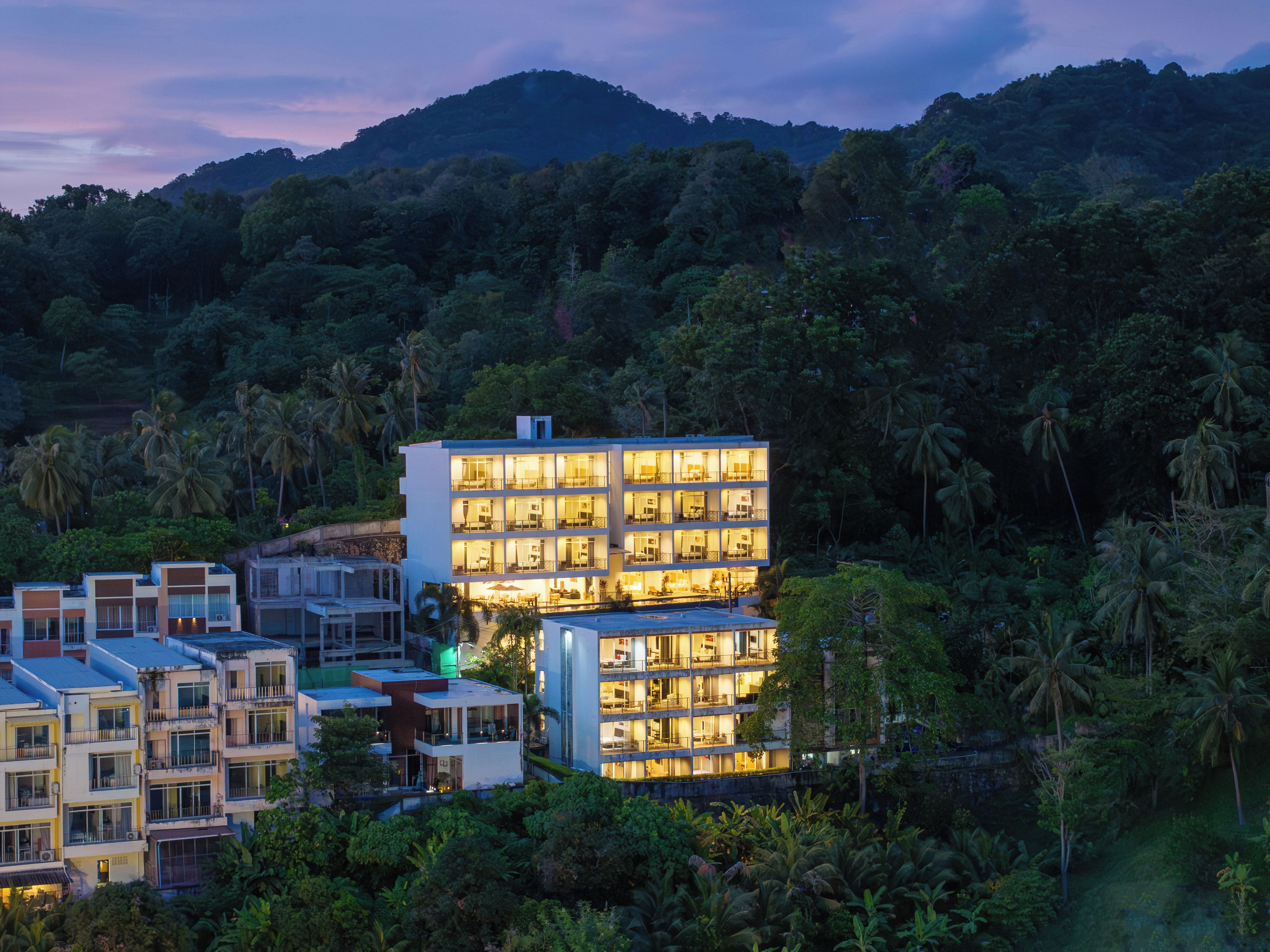 A multi-story hotel with balconies, surrounded by lush greenery and hills.