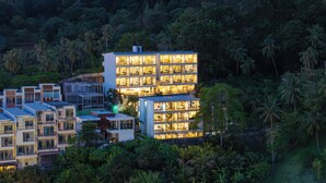 Un hotel de varios pisos con balcones, rodeado de abundante vegetación y colinas.