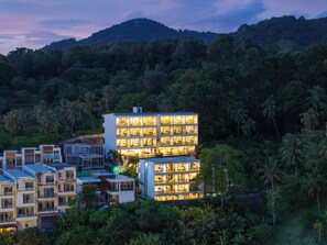 Un hotel de varios pisos con balcones, rodeado de abundante vegetación y colinas.