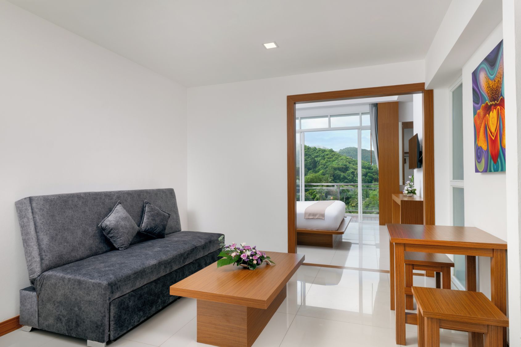 Una habitación de hotel moderna con un sofá gris, una mesa de centro de madera, una cama con vistas y un cuadro en la pared.
